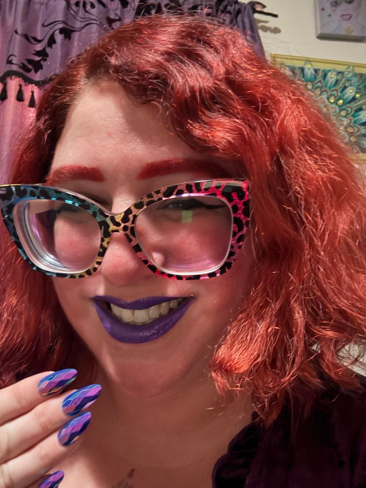 A picture of Stephanie smiling at the camera. She has chin length red hair, matching eyebrows, big glittery square rainbow leopard glasses, and purple lips. She is holding one hand by her chin with medium length almond shaped 3D geometric shaped nails. One side of her purple velvet dress can be seen.