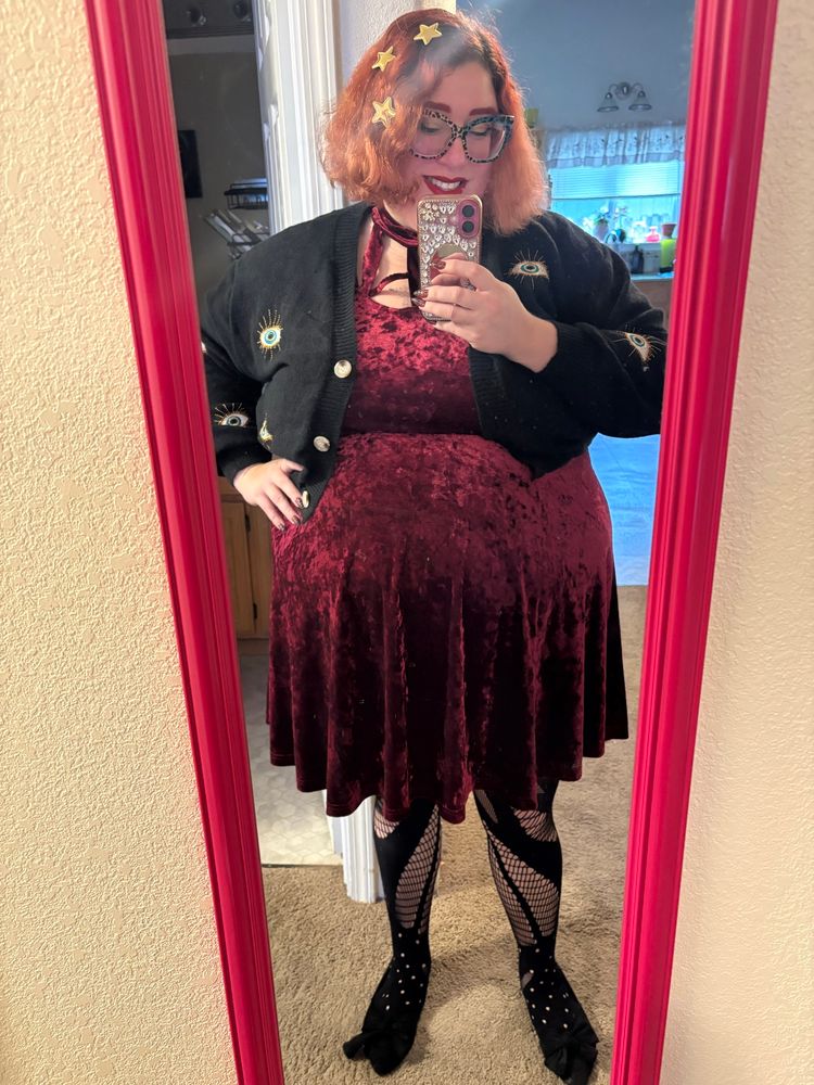 A full body mirror selfie of Stephanie smiling down at her bejeweled pink phone. She has chin length wavy red hair with gold stars in it, red eyebrows, big glittery square rainbow leopard glasses, and red lips. She is wearing a cropped black knit sweater embroidered with blue and gold evil eyes over a burgundy velvet dress over patterned black tights and pointy black shoes. She is standing in a hallway in front of a kitchen.
