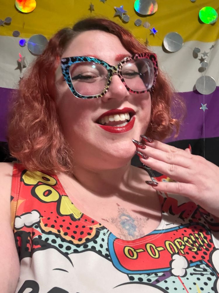 A picture of Stephanie smiling down at the camera in front of a nonbinary flag with circle and star garlands hung aesthetically around. Stephanie has chin length wavy red hair, matching eyebrows, but glittery square rainbow leopard glasses, and red lips. She has one hand at her chin with medium length almond shaped red nails with black designs on them. She is wearing a red, yellow, blue, and black dress with big comic book POW and BANG action bubbles and clouds on it.