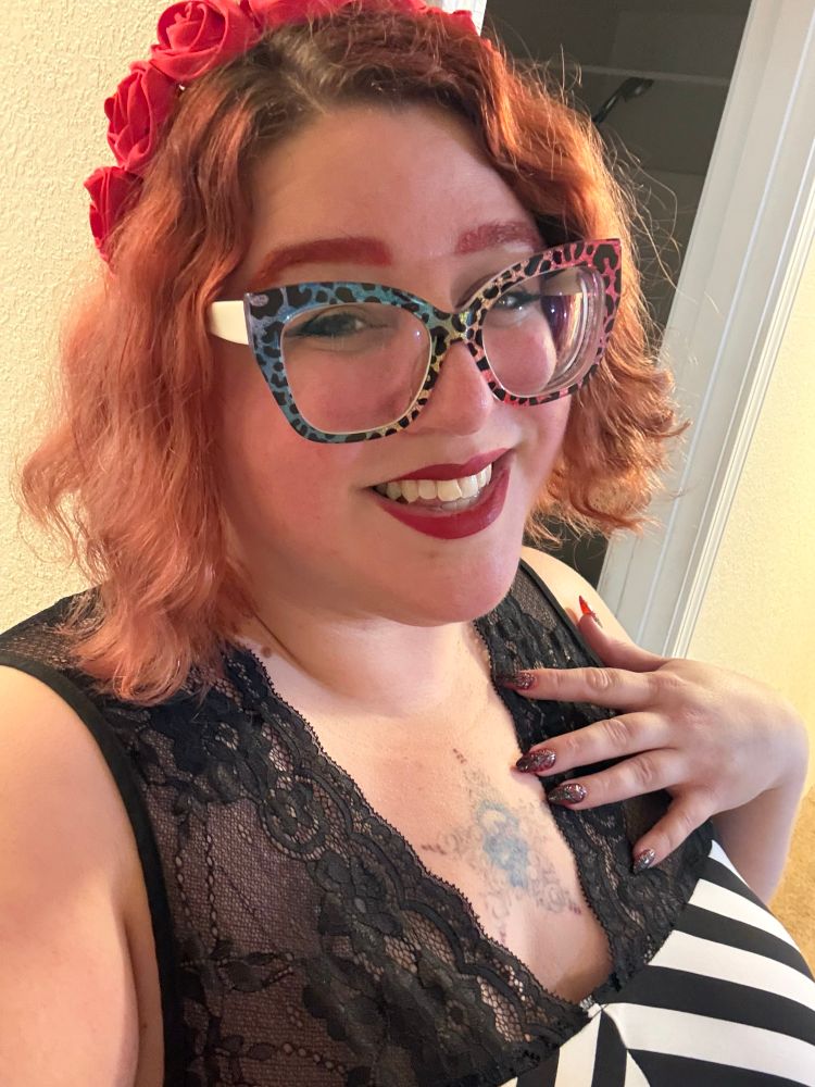 A picture of Stephanie smiling at the camera. She has chin length wavy red hair with a headband of red silk roses on, red eyebrows, big glittery square rainbow leopard glasses, and red lips. She is wearing a black and white striped dress with thick black lace straps. She has one hand against her chest with medium length almond shaped red nails with black designs on them.