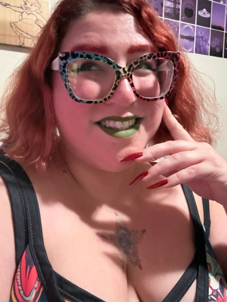 A picture of Stephanie smiling and looking off camera. She has medium length wavy red hair that falls to her chin, matching eyebrows, big glittery square rainbow leopard glasses, and green lips. She has one hand against her face with long red stiletto nails on it. She is wearing a dress with two black straps on either shoulder and angry cat faces on it.