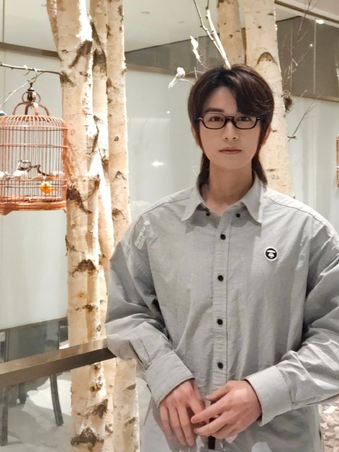 Chinese actor Zhu Zanjin stands next to a songbird cage that hangs from a tree. he is wearing a pale oversized dress shirt that harmonizes with the light background, and has black glasses and grown-out hair. his stance is casually elegant. note: we haven't seen his hair this long for years.