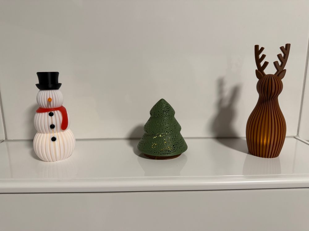 Drei Figuren aus dem 3D-Drucker.

Ganz links ein Schneemann mit rotem Schal, orangener Nase, schwarzem Hut & Knöpfen.

In der Mitte ein kleiner dunkelgrüner Weihnachtsbaum.

Rechts ein minimalistisches Rentier in braun. 