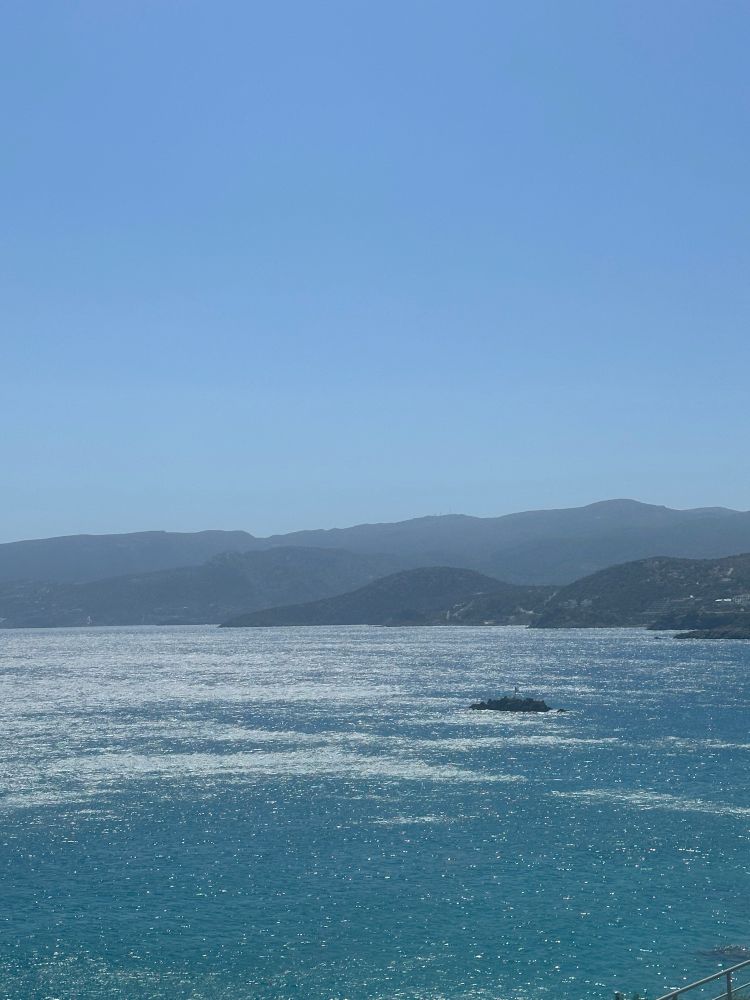 Blaues, gekräuseltes Meer im Vordergrund, Felseneinschluss im Meer, im Hintergrund Gebirgssilhouette, blauer Himmel.