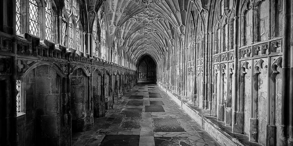 A long stone corridor with a vaulted Gothic ceiling, viewed in sharp perspective. Intricate tracery arches line the left side, casting shadows across the patterned floor. The corridor narrows toward a dark, closed doorway in the distance. The image is in black and white, emphasizing the depth, symmetry, and aged texture of the stone.