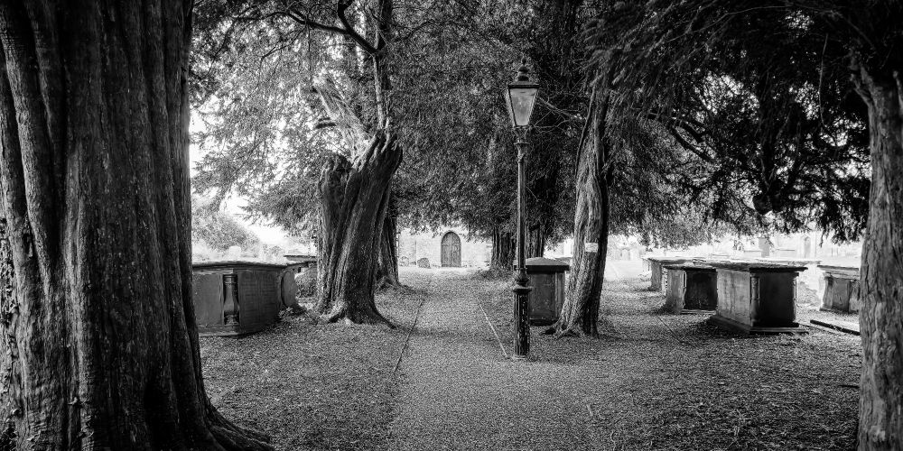 A narrow gravel path leads through a graveyard toward a wooden church door in the village of Tormarton, UK. Tall yew trees and weathered tombs line the path on either side. A gas lamp stands near the center, and the trees’ branches form a dark canopy overhead. The image is in black and white, emphasizing depth, texture, and quiet symmetry.