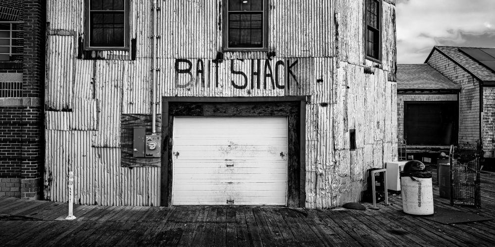 Black-and-white photograph of an old corrugated-metal building labeled “Bait Shack.” A closed garage door sits beneath hand-painted lettering, flanked by weathered siding and small industrial objects—trash can, lobster trap, and cables. The rough wood planks of the dock lead toward the structure, whose worn textures evoke age, labor, and the quiet endurance of a working harbor in the off-season.
