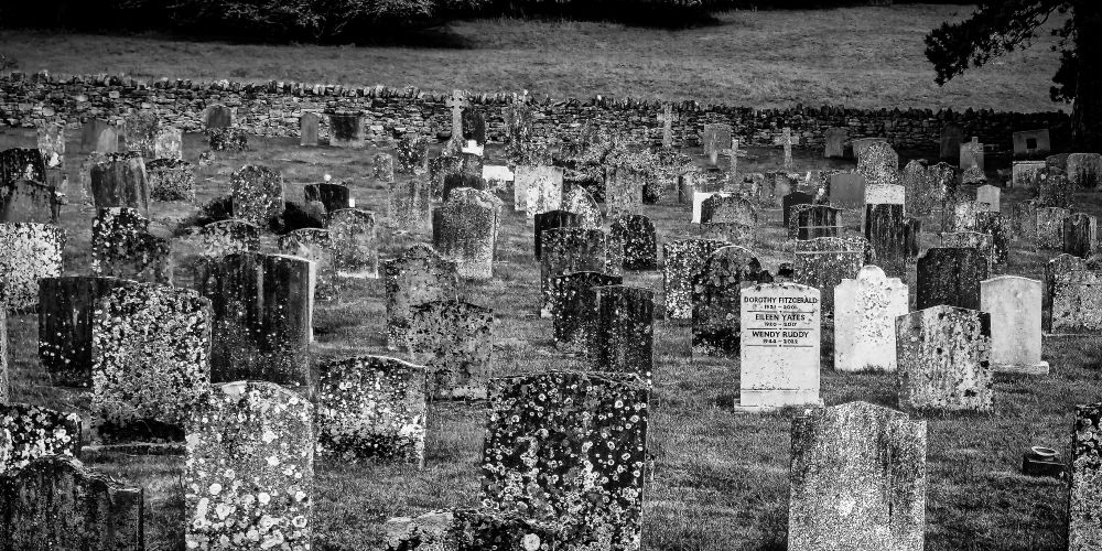 Black and white photo of a rural church graveyard with many, many mossy covered gravestones, with a single modern gravestone in their midst.