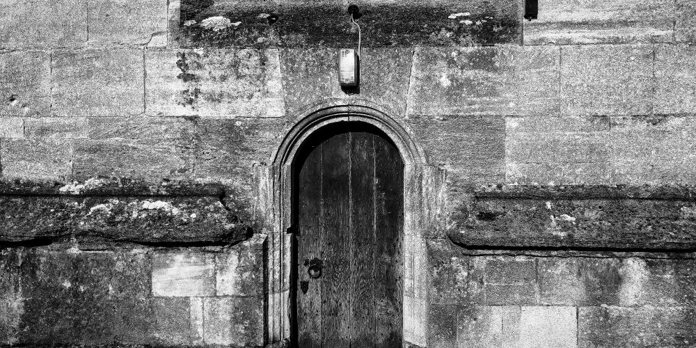 A weathered wooden church door sits within a rounded stone arch, centered on a large wall of aged limestone blocks. Above the door, a rectangular exterior light fixture is mounted on a cable. The stonework is mottled and textured, showing erosion and discoloration. The image is in black and white, emphasizing symmetry, stone texture, and the quiet contrast between architecture and utility.
