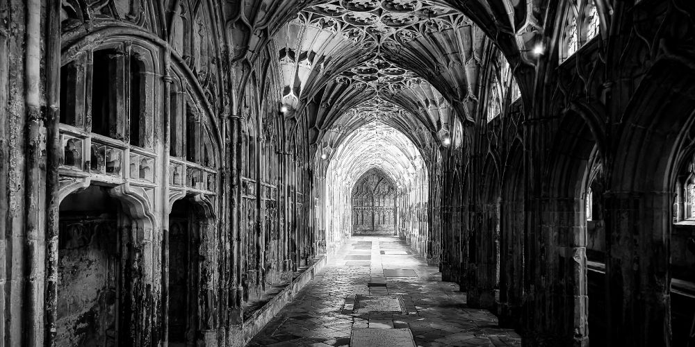 A black and white image of the vaulted stone cloisters of Gloucester Cathedral stretching into the distance, with intricate tracery overhead and worn floor slabs leading to a bright archway at the far end.