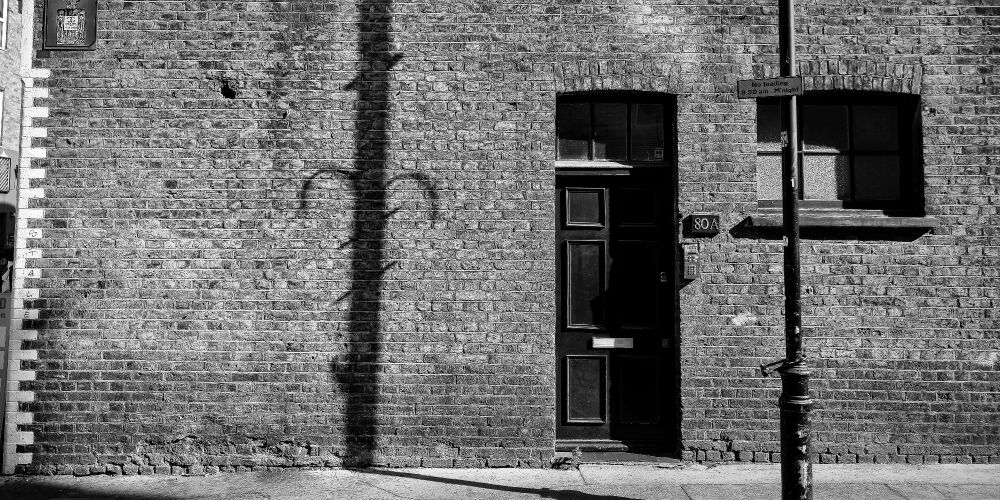 A black and white image of a lamppost casting a shadow on a large blank  brick wall, next to a door for a residence with the house number 80A.  The brick wall is in Covent Garden, UK