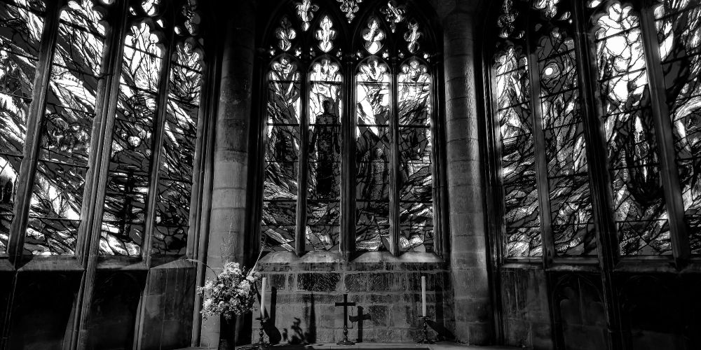 A black and white image of the blue stained glass window in Gloucester Cathedral fills the upper frame, its swirling designs casting dramatic light. Below, a stone altar holds two candleholders and a cross, with their shadows falling onto the wall. A bouquet of flowers stands to the side. The image is in black and white, emphasizing structure, reflection, and sacred stillness.
