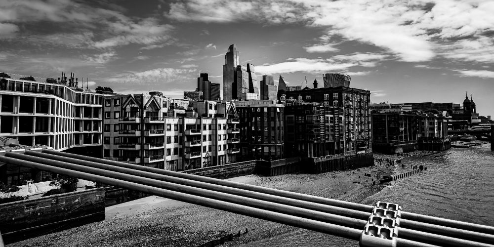 A black-and-white photograph taken from the Millennium Bridge in London. Steel support pipes cross the foreground, leading toward a layered cityscape of residential flats, brick warehouses, and modern glass towers. The River Thames curves along the lower right, and the skyline rises beneath a sky of broken clouds.