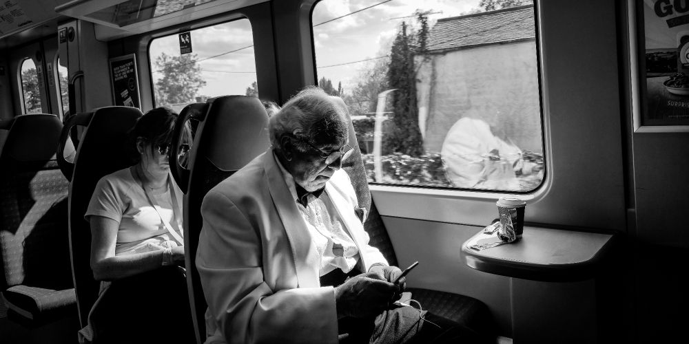 A man in a white tuxedo sits alone in a train carriage, his head slightly bowed as he looks at his phone. Soft light from the window illuminates his jacket, creating a stark contrast with the darker seats and shadows around him. Another passenger, partially visible beside him, adds subtle human context. The scene is quiet, reflective, and formal, rendered in black and white.