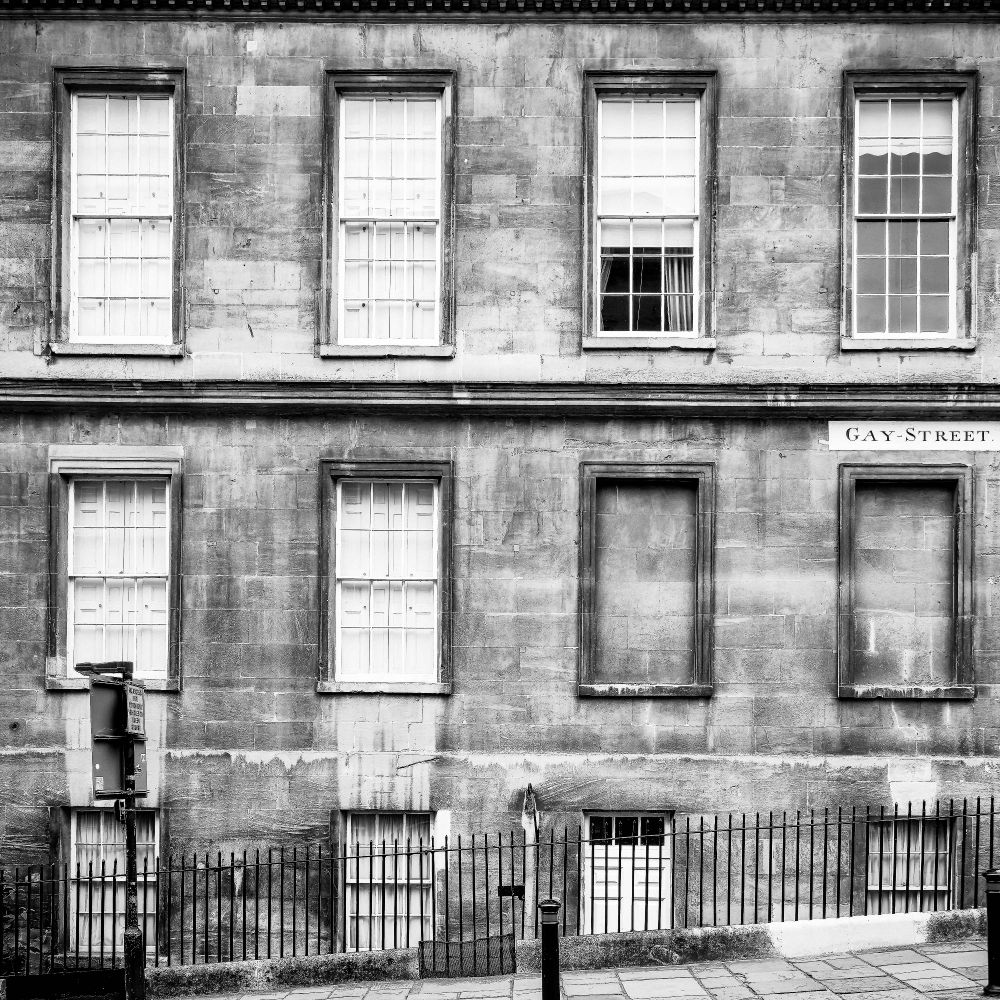 Black and white image of three stories of windows in a building along Gay Street in Bath, UK.  One row of the windows lies along and below street level, and a low metal fence protects pedestrians along the sidewalk from falling into the void immediately in front of the windows.  The windows vary in what is visible from the street.  A sign reading Gay Street is attached to the building.