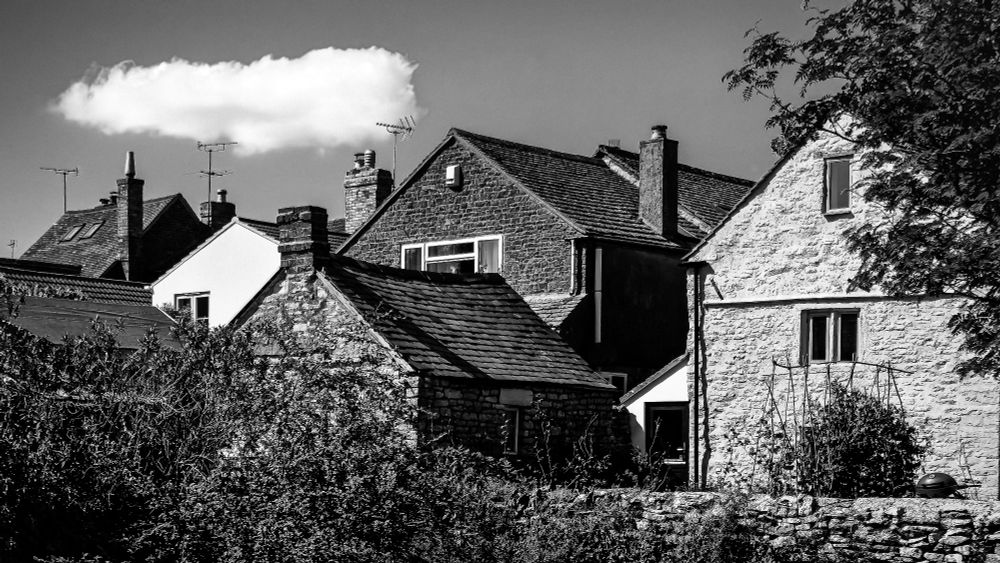 Black and white photo of several rooftops, their pointed tops in harmony.  Above them, a lone, small white cloud floats above the rooftops.