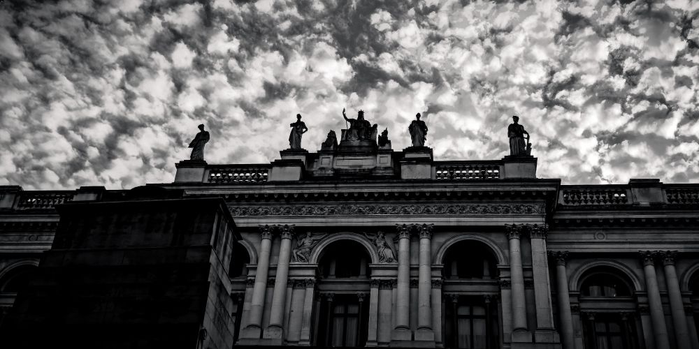 A row of stone statues stands atop a neoclassical government building in Whitehall, London. The figures are silhouetted sharply against a sky filled with rippling, textured clouds, creating a dramatic contrast between the dark architectural forms and the illuminated sky. The photograph is in black and white, emphasizing shape, line, and shadow.