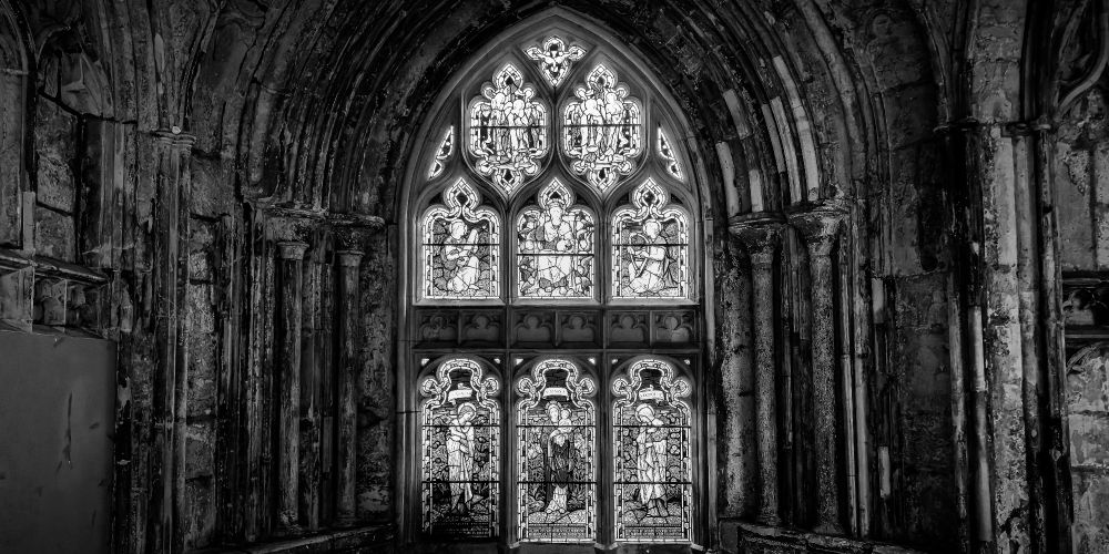 A tall Gothic stained glass window set within a deeply shadowed stone archway. The window is composed of ornate panels, with detailed tracery at the top and six religious figures below. The surrounding stonework is aged and textured, with worn pillars and crumbling surfaces framing the illuminated glass. The image is in black and white, emphasizing the contrast between light and decay, detail and darkness.