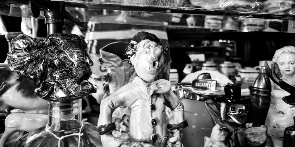 A close-up of a group of ceramic and porcelain figurines arranged tightly on a shelf. In the center is a clown-like figure with exaggerated features and a wide-eyed expression. Surrounding it are a shiny elephant bust, a parrot, and various other whimsical or surreal objects, all reflected in the glass above and behind them. The photo is black and white, emphasizing texture and visual clutter.
