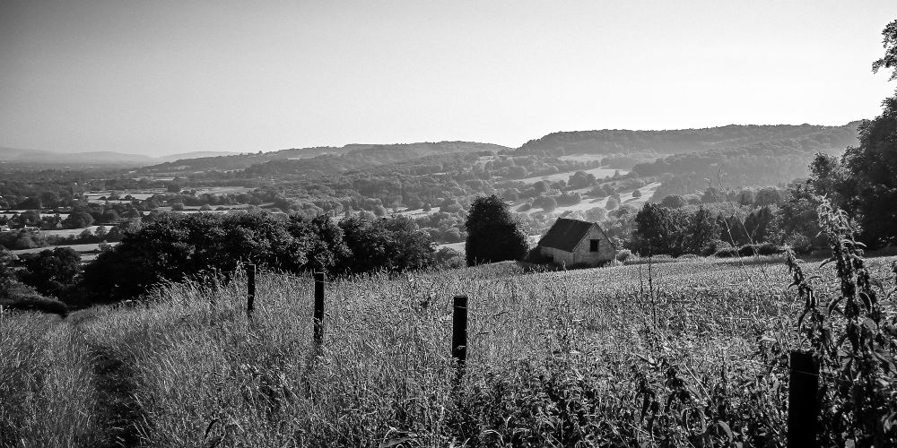 Black and white image of a barn on a hillside field, with hills and the escarpment of the Cotswolds extending off into the background.