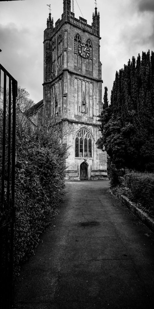 A black-and-white photograph of St. James the Great Church in Dursley, England. Viewed from the center of a paved path, the stone tower rises above an arched entrance, flanked by hedges and iron gates. A large clock is mounted on the tower, and the overcast sky enhances the mood of stillness.