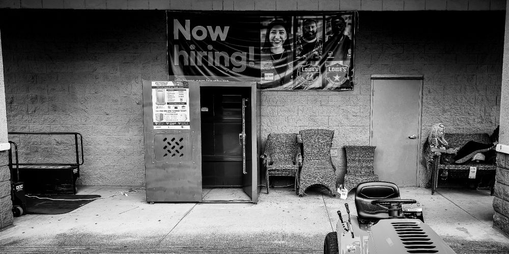 A black-and-white photo of the exterior of a Lowe’s hardware store. A large “Now hiring!” banner hangs above a metal locker, wicker furniture, and a closed door. A woman sits on a patio couch to the right, legs crossed, absorbed in her phone. A riding lawnmower and price tags are visible in the foreground. The image captures irony and stillness in a commercial setting.