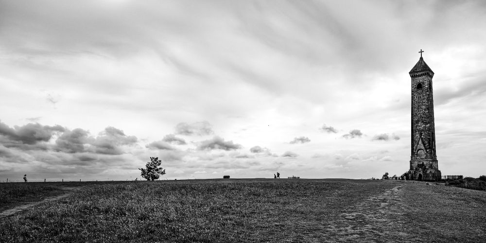 The Tyndale Monument in the Cotswolds rises on the right side of the frame beneath a sweeping sky, with a small group of walkers and a bench dotting the distant horizon. The ground slopes gently upward, covered in wild grass and a faint footpath leading into the scene. The image is black and white, emphasizing contrast and stillness.

