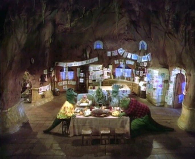 A very wide shot of the set and cast of the 90s television show Dinosaurs. The interior scene sits in the shell of a volcano and the family sits at the dining room table happily eating dinner. Along the back wall are various doors and the kitchen of the house. The photo quality is not great. The refrigerator seems to be covered fully in papers, but whether they're drawings, bills, letters cannot be determined. The floor is a square, brick like pattern. Generally earth tones with greige appliances and accents. 