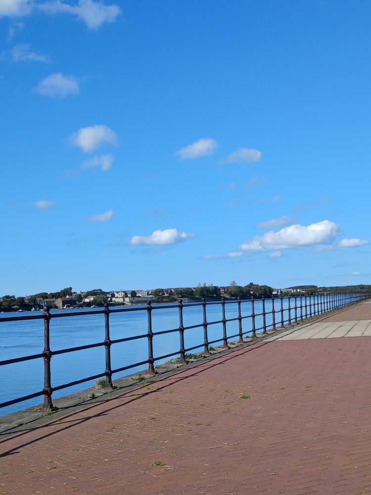 Red brick promenade, railings, bright blue sea in the Walney channel and house in the distance on Walney island under a blue sky with small white scudding clouds.
