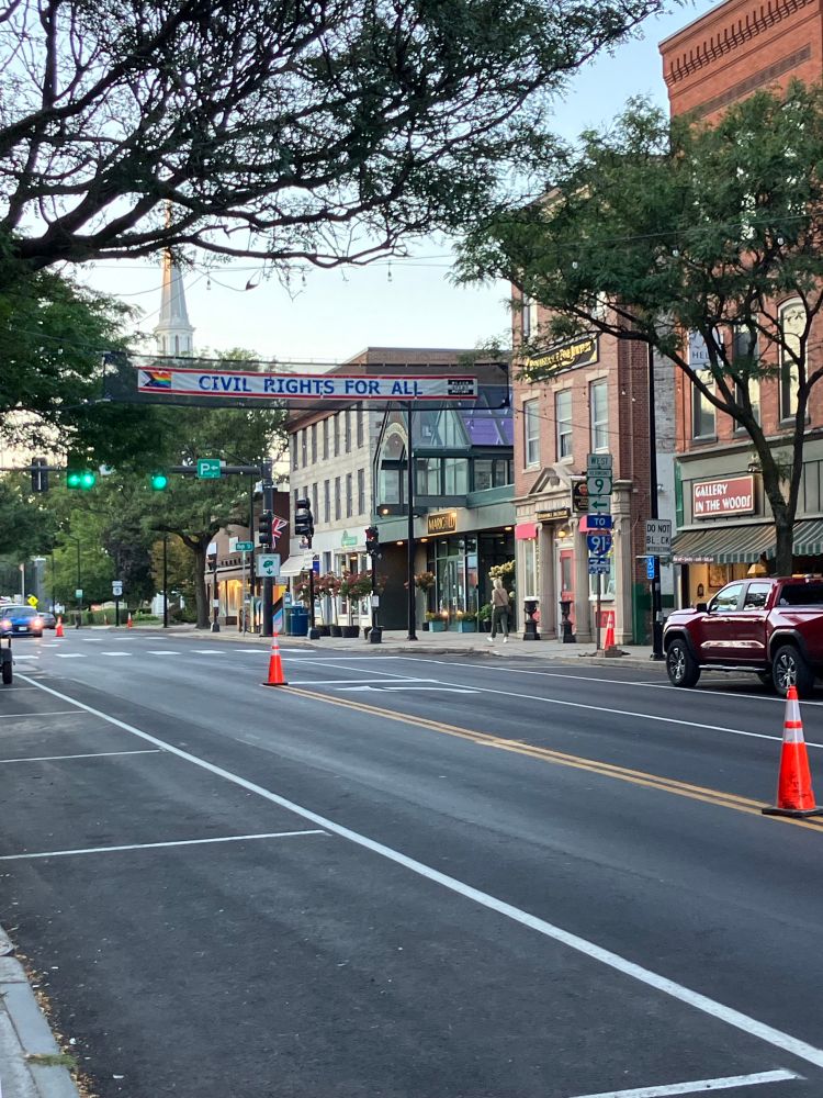 Downtown Brattleboro, Vermont. There is a sign hung above the road that reads “Civil rights for all” with a pride flag on one side and a Black Lives Matter sign on the other. The road stretches into the distance and there are storefronts along the right side. There are some trees on the other side of the road. 