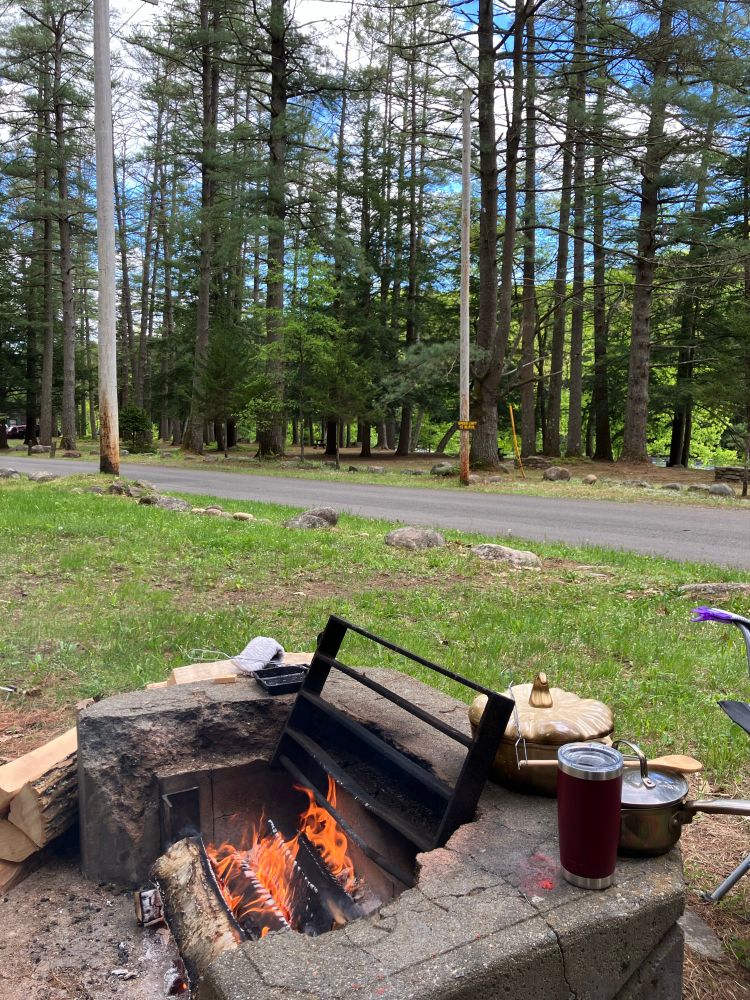 A picture of a campsite. At the bottom of the picture there is a concrete fire pits with a fire going in it and various cooking pots, cups, and logs around it. In the background there is some grass, a road, and then a wooded area with tall pine trees. The sky is blue with some clouds. 
