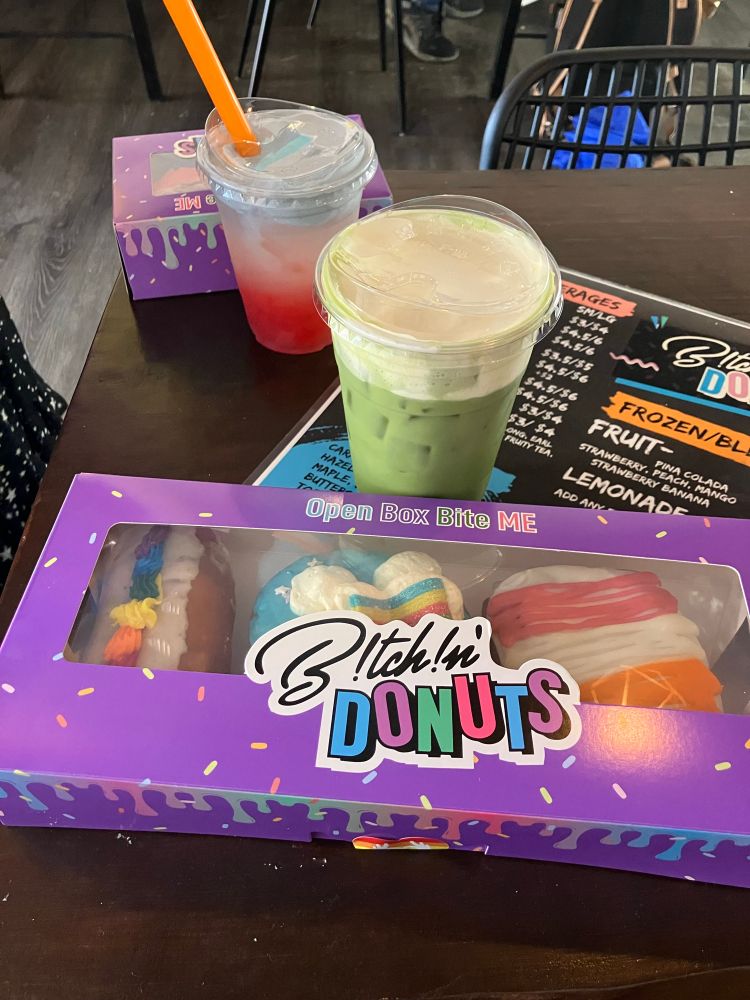 A picture of 3 pride themed donuts (one frosted rainbow colored, one blue with white clouds and a rainbow candy, and one frosted pink, white, and orange like a lesbian flag) in a purple box with sprinkles and the Bitchin’ Donuts logo on it. There is a green matcha drink behind it. There is another donut box and colorful drink in the background. 