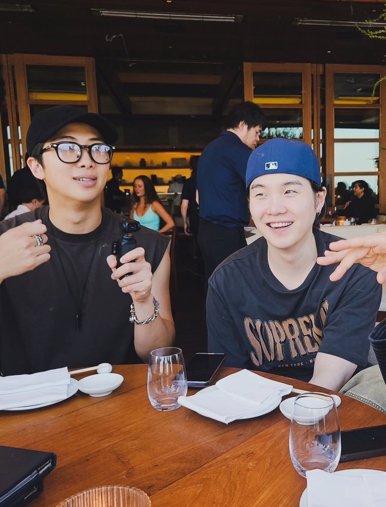 Photo of Namjoon and Yoongi sitting at the table. Namjoon is wearing black sleeveless shirt and a cap. Yoongi is also wearing black shirt and blue cap. They look so cute