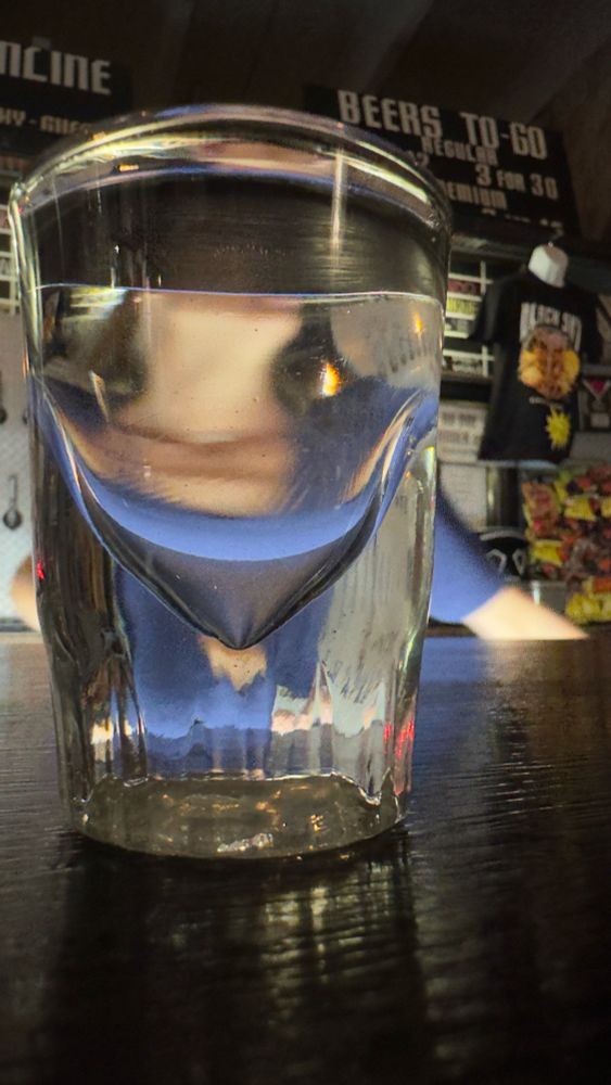 A shot glass filled with clear liquor that is distorting a woman behind the bar