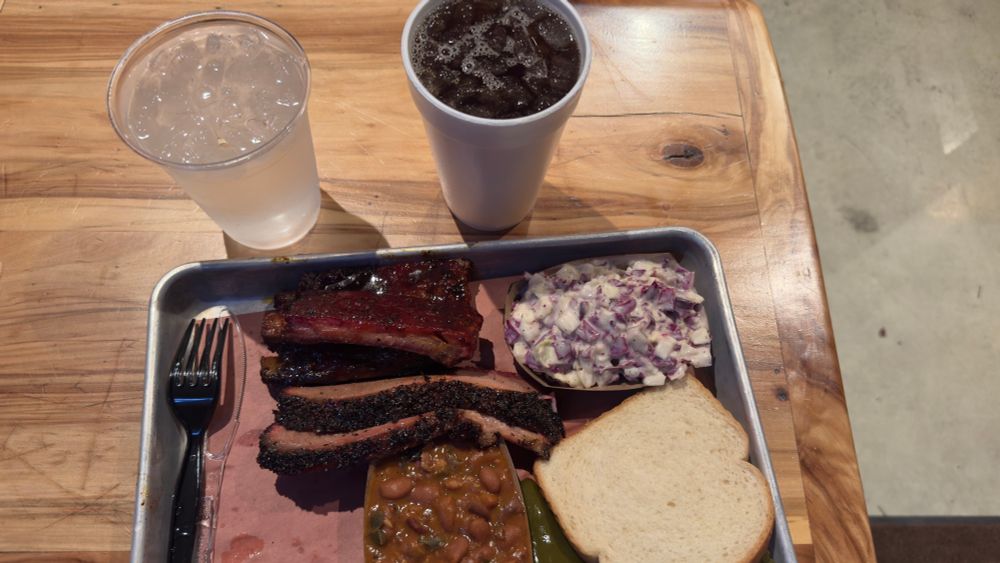 A steel plate with brisket, ribs, beans, slaw, white bread, pickles, onions, and jalapeños along with a sweet tea and water