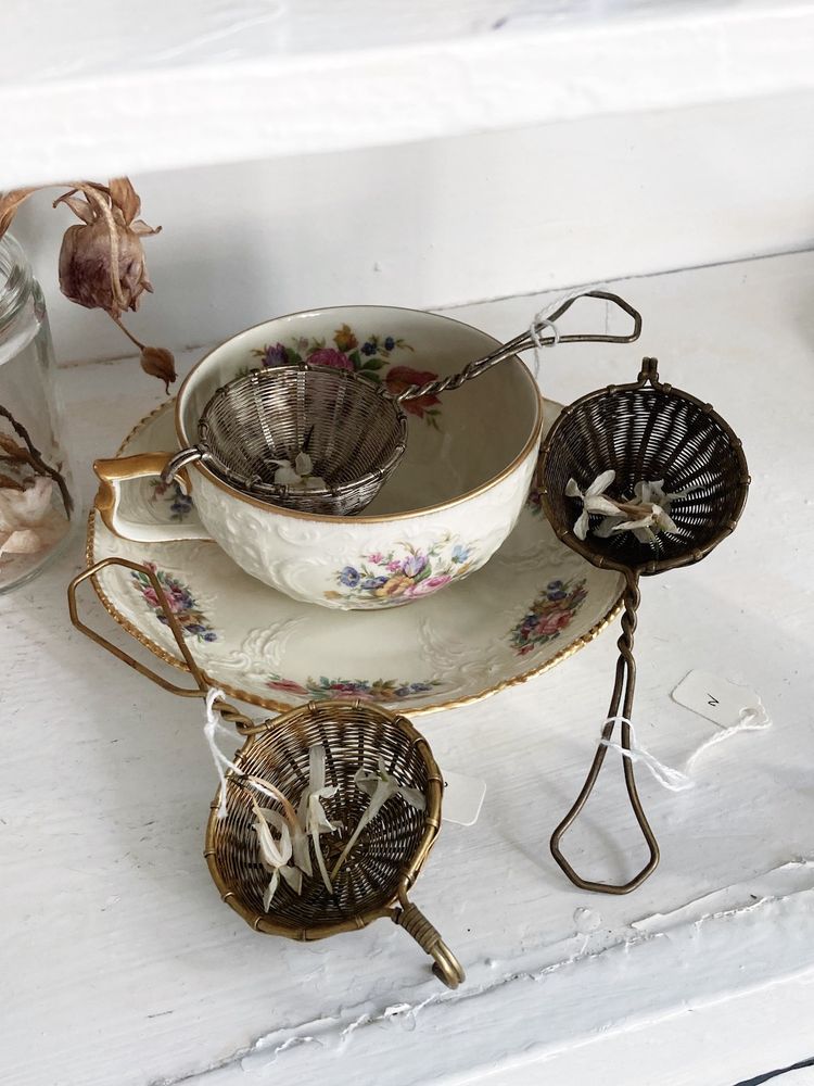 A vintage teacup and saucer with floral pattern, surrounded by three small mesh tea strainers with twisted metal handles. Small white flower petals are visible inside the strainers. The arrangement sits on a distressed white wooden surface, with a dried flower visible in the background.