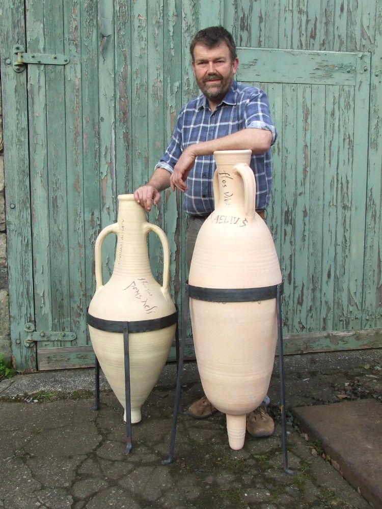 Graham Taylor of Potted History, standing behind two of his replica Roman Amphorae. He is resting his elbow on the rim of the larger of the two which stands at one point two metres  tall,