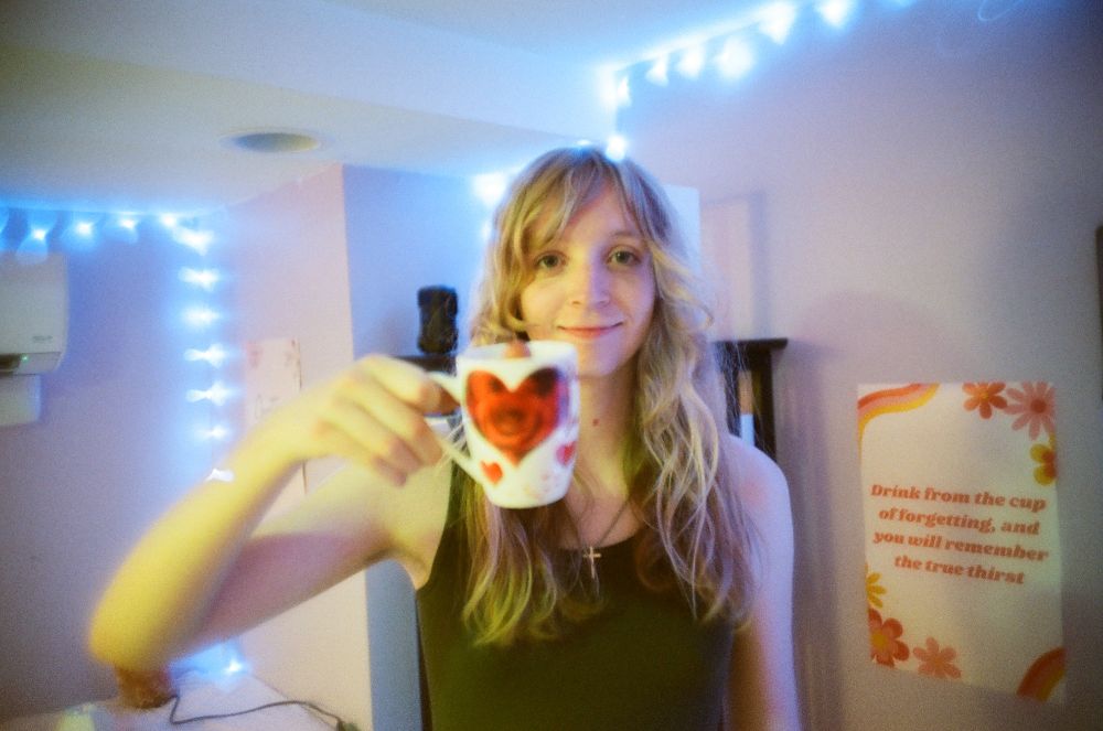 Lex Walton poses with a coffee mug that has a heart on it and contains a hot dog. Next to him is a poster which reads "Drink from the cup of forgetting and you will remember the true thirst."