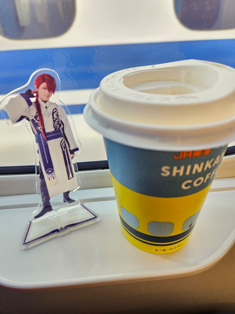 新幹線の窓際の小さなテーブルの上。右にドクターイエローのイラストが入った蓋つきの紙コップ、中身はコーンスープ。左に今回観劇しに行くまほステのグッズのカインのアクリルスタンドが置かれている。窓の奥には白い車体に青いラインが入った車体が見えている。