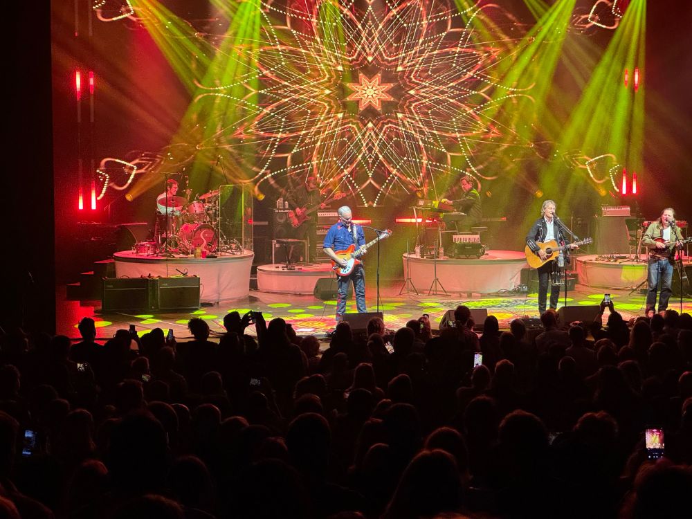 view over the crowd in shadow, 
drummer to the left, bass player, organist, guitar player at the back of the stage
three singer/guitar players at the front of the stage
yellow / orange lighting with complex geometric design on the back screen
