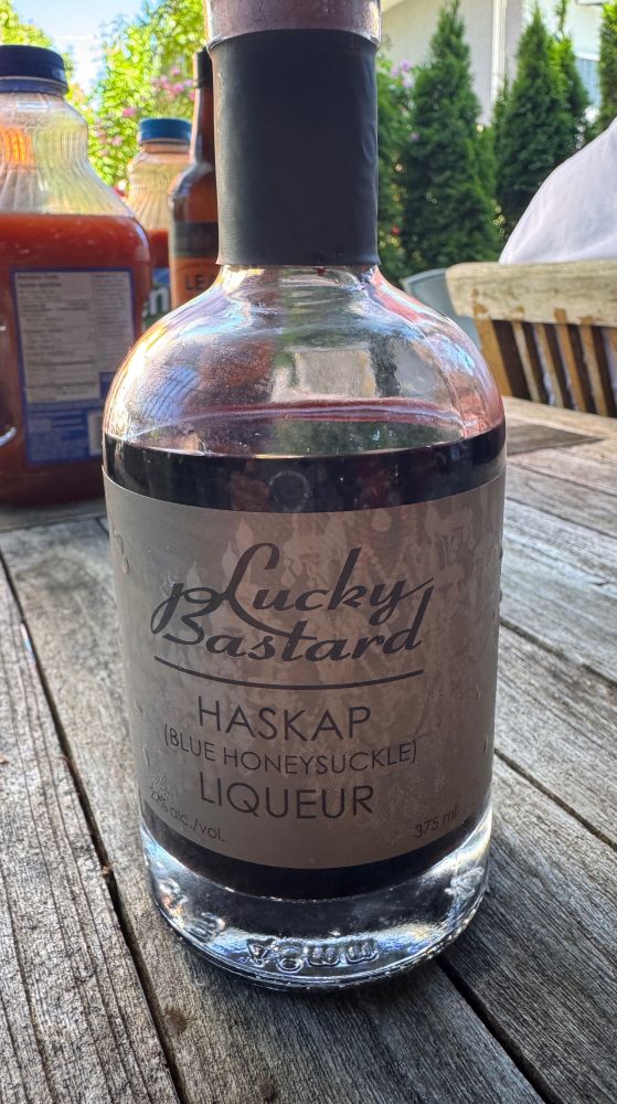 bottle with dark purple liquid sitting on weathered wooden table. Label says: 
Lucky Bastard 
Haskap
(Blue Honeysuckle)
Liqueur