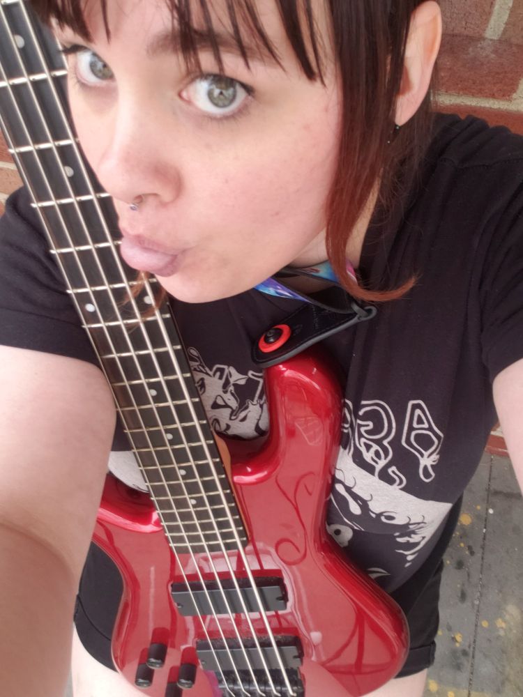 An Alice posing with a red 5 string bass on her lap and her tongue out