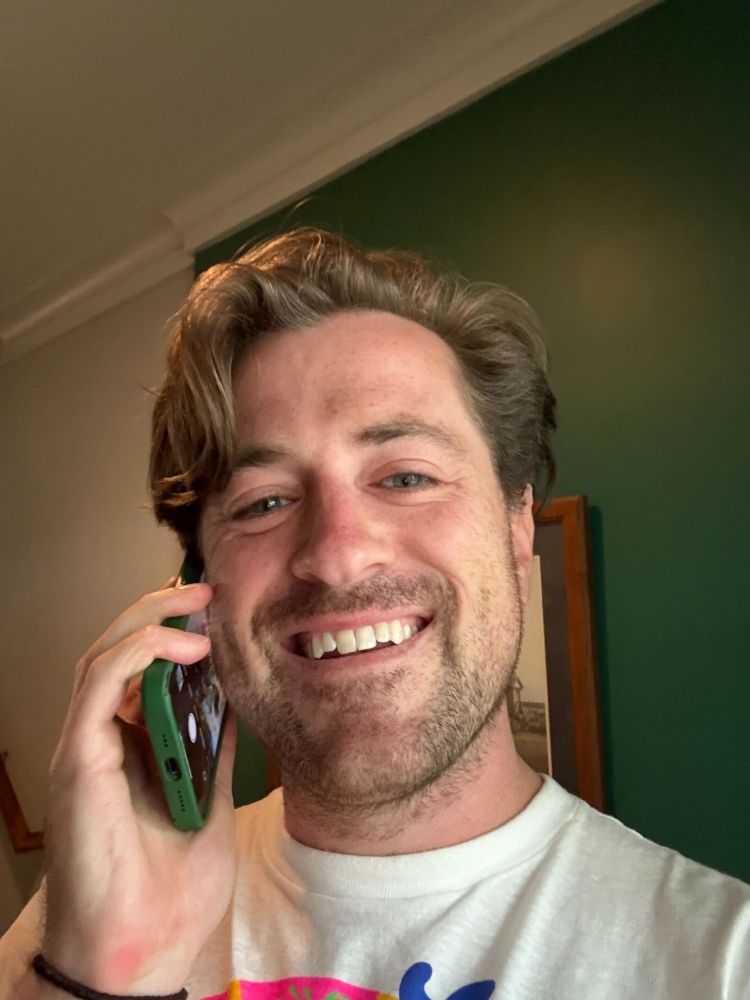 A man with brown hair smiling big on the phone
