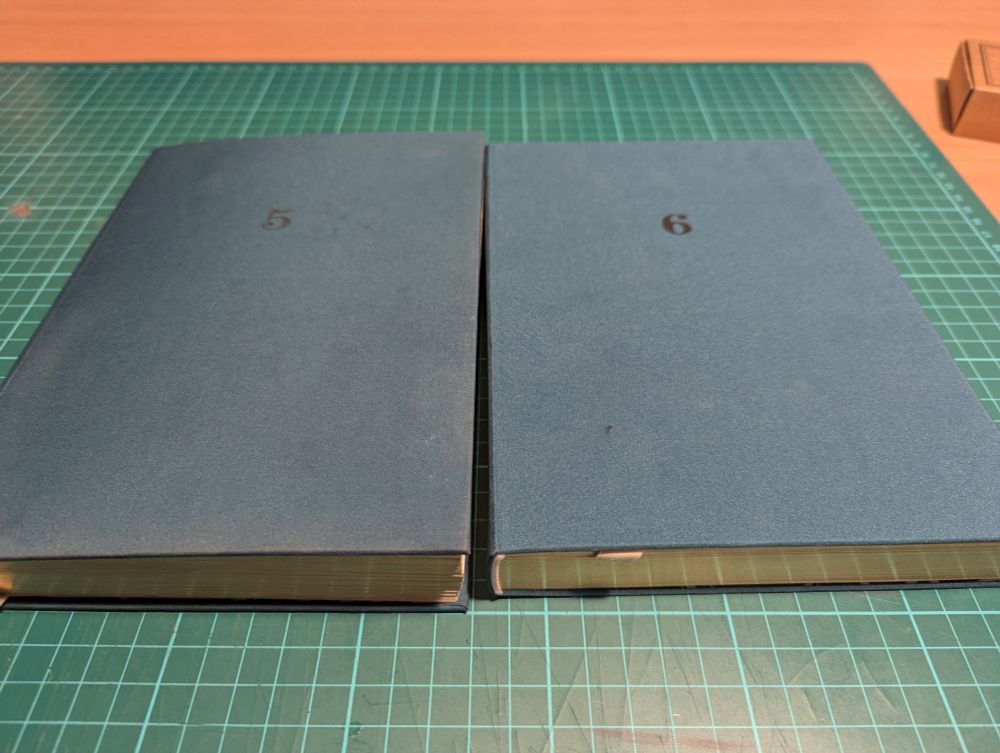 To identical journals side by side. One is a but old and battered and has the number 5 printed on it. The other is very shiny and new with the number 6 printed on the front. They are lying on a green cutting mat.