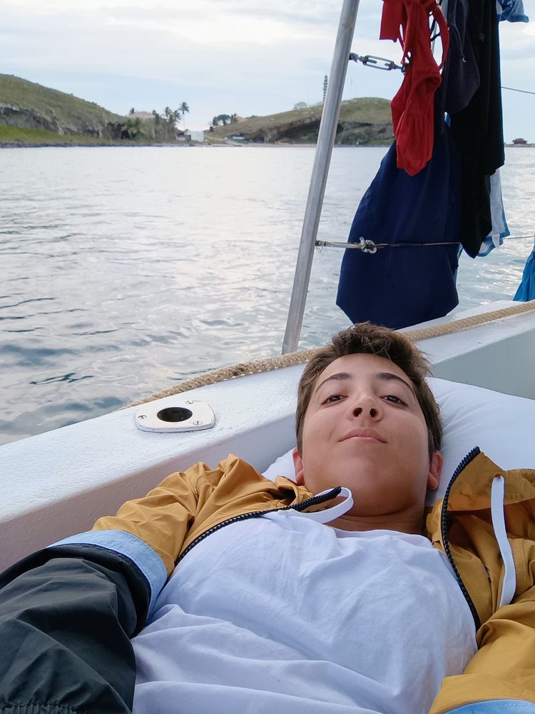 Eu, mulher branca de cabelo curto, deitada num travesseiro branco em um barco. Ao fundo é possível ver o mar