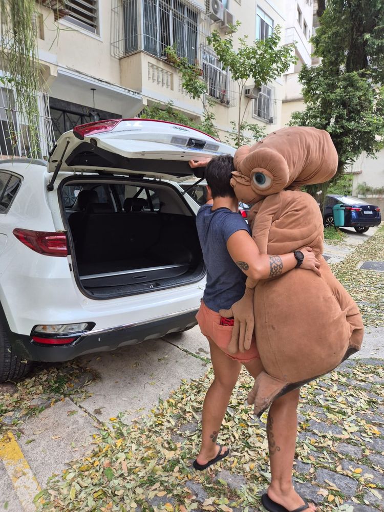 Eu, uma mulher branca de cabelo curto magra, usando short laranja e camisa azul, fechando a mala de um carro branco e segurando um boneco gigante do ET do Spielberg. O ET está com a mão na minha bunda 