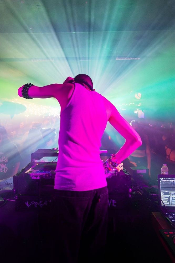 A photo of a DJ from behind, djing to a barely visible crowd in a foggy room. Beams of light point out from the center of the frame