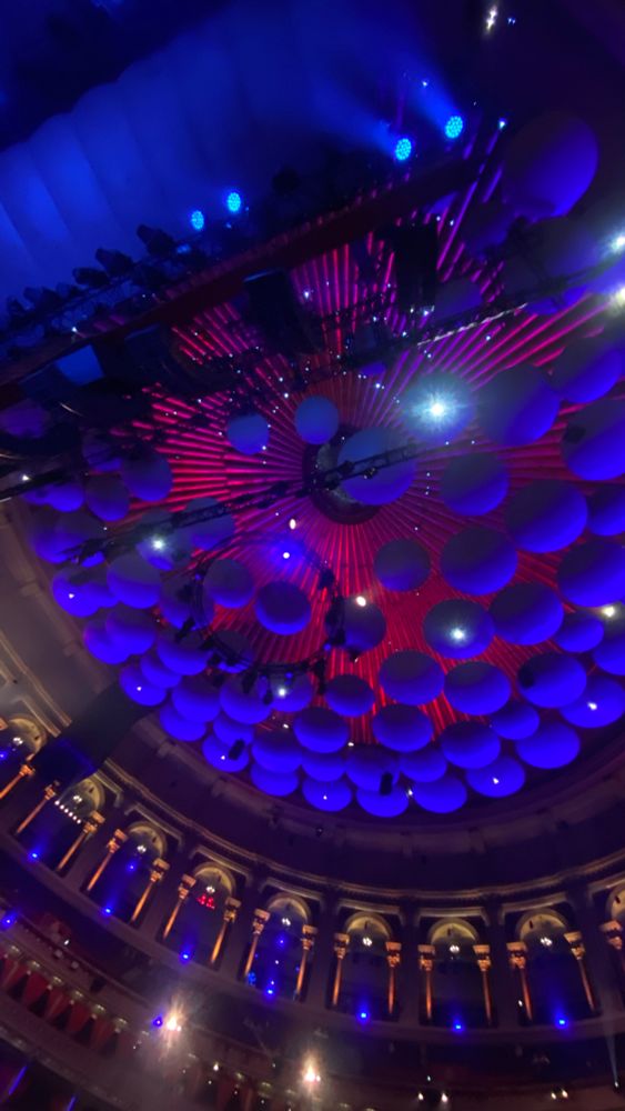 Looking up to the “acoustic mushrooms” hung below the roof of the Albert Hall to reduce echo, lit purple with roof redder. Some of the arches around the Gallery level are also visible 