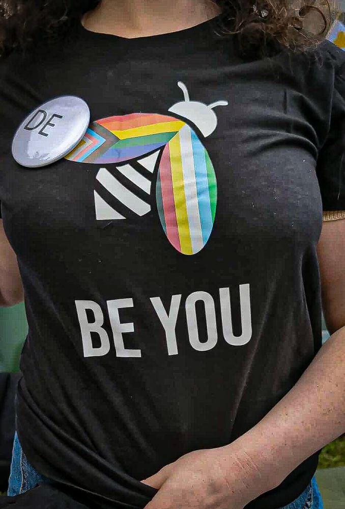 Svart t-skjorte med et trykk av en stilisert bie med inklusive intersex Prideflagget på venstre vinge og funkisPride-flagget på høyre. Under står i store hvite bokstaver BE YOU.
Personen som har på t-skjorten holder den ned slik at vi ser trykket bedre, og har en stor hvit pin som sier DE