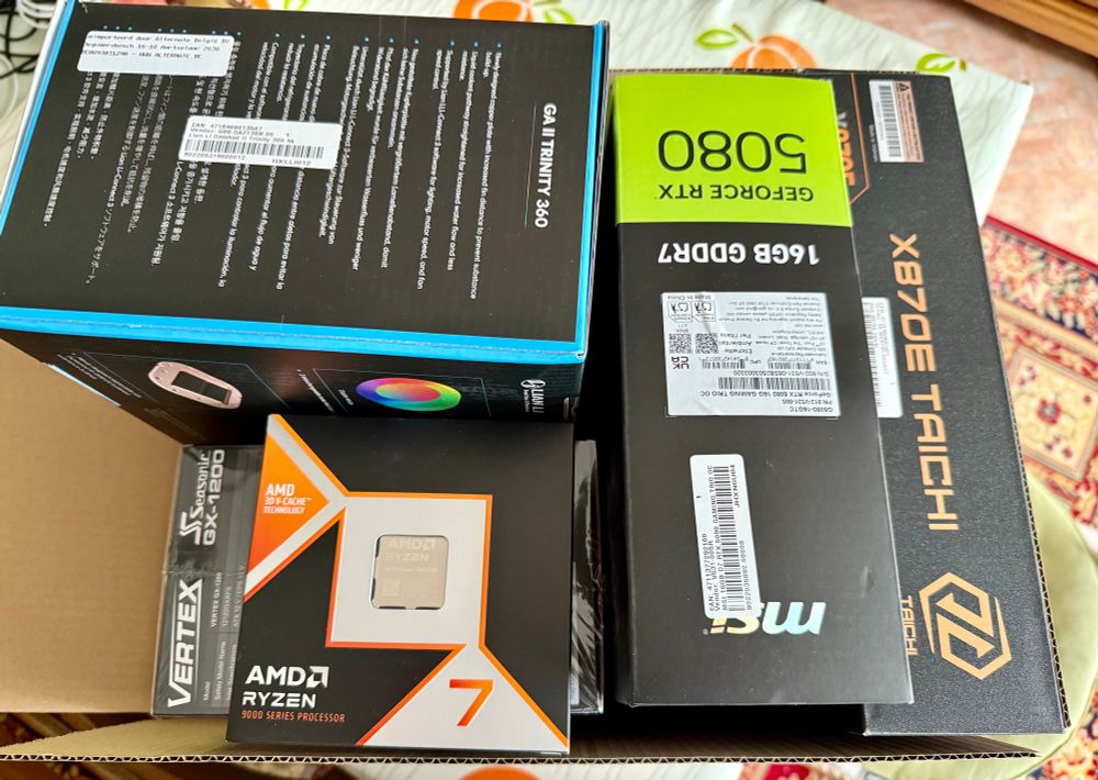 Boxed PC parts, showing an AMD Ryzen 9800X3D, MSI GeForce RTX 5080, Asrock X870E Taichi motherboard, AIO watercooling and a power supply 